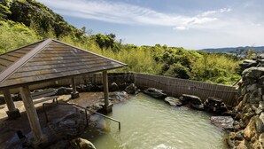 Perinteinen japanilainen onsen-kylpylä, jossa on puinen paviljonki, kivinen kylpyallas ja taustalla kivikkoinen maisema.