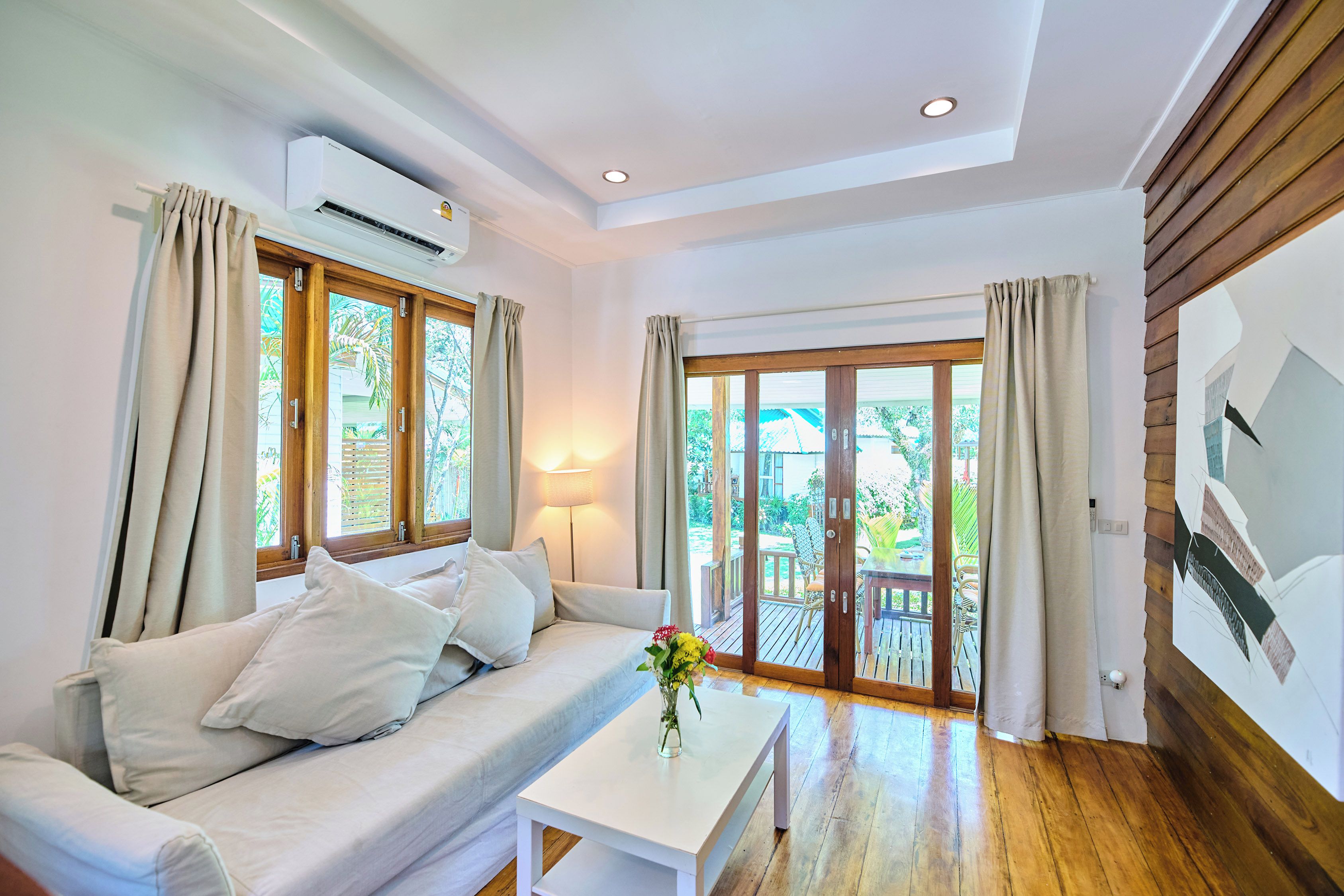 A modern living room with a white sofa, a small table with a vase, and a view of a wooden deck and greenery outside.