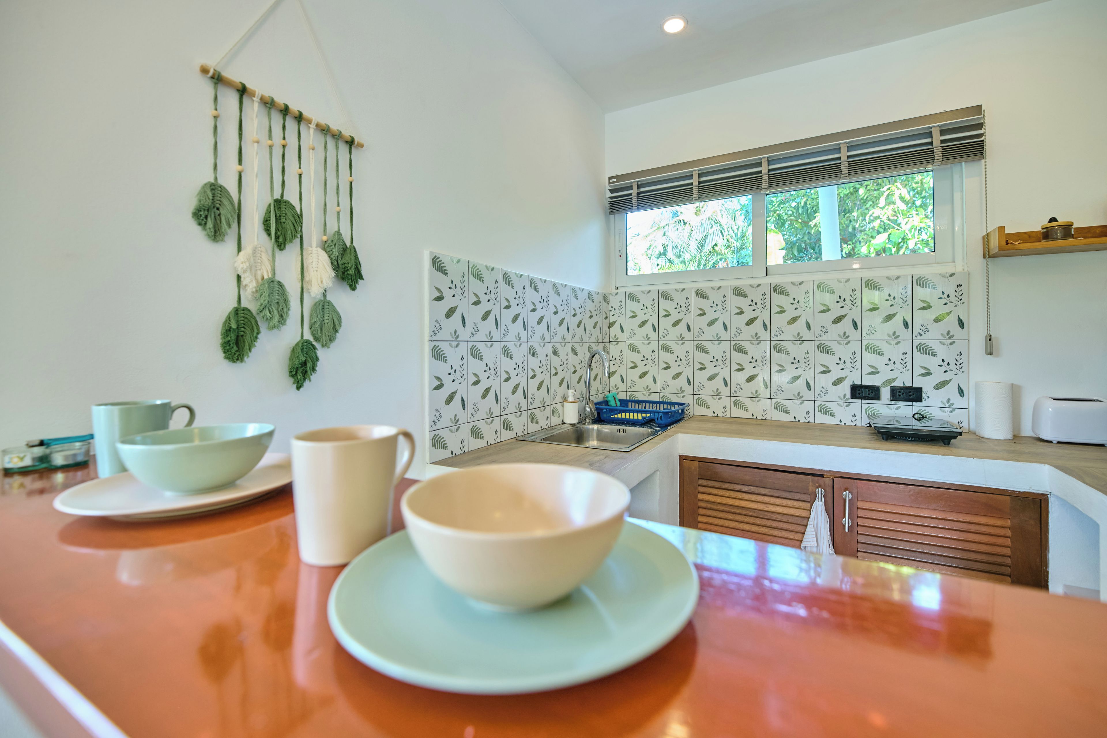 Una cocina con encimera de madera, una planta decorativa colgante, una ventana con persianas y un salpicadero de azulejos con diseño de hojas.