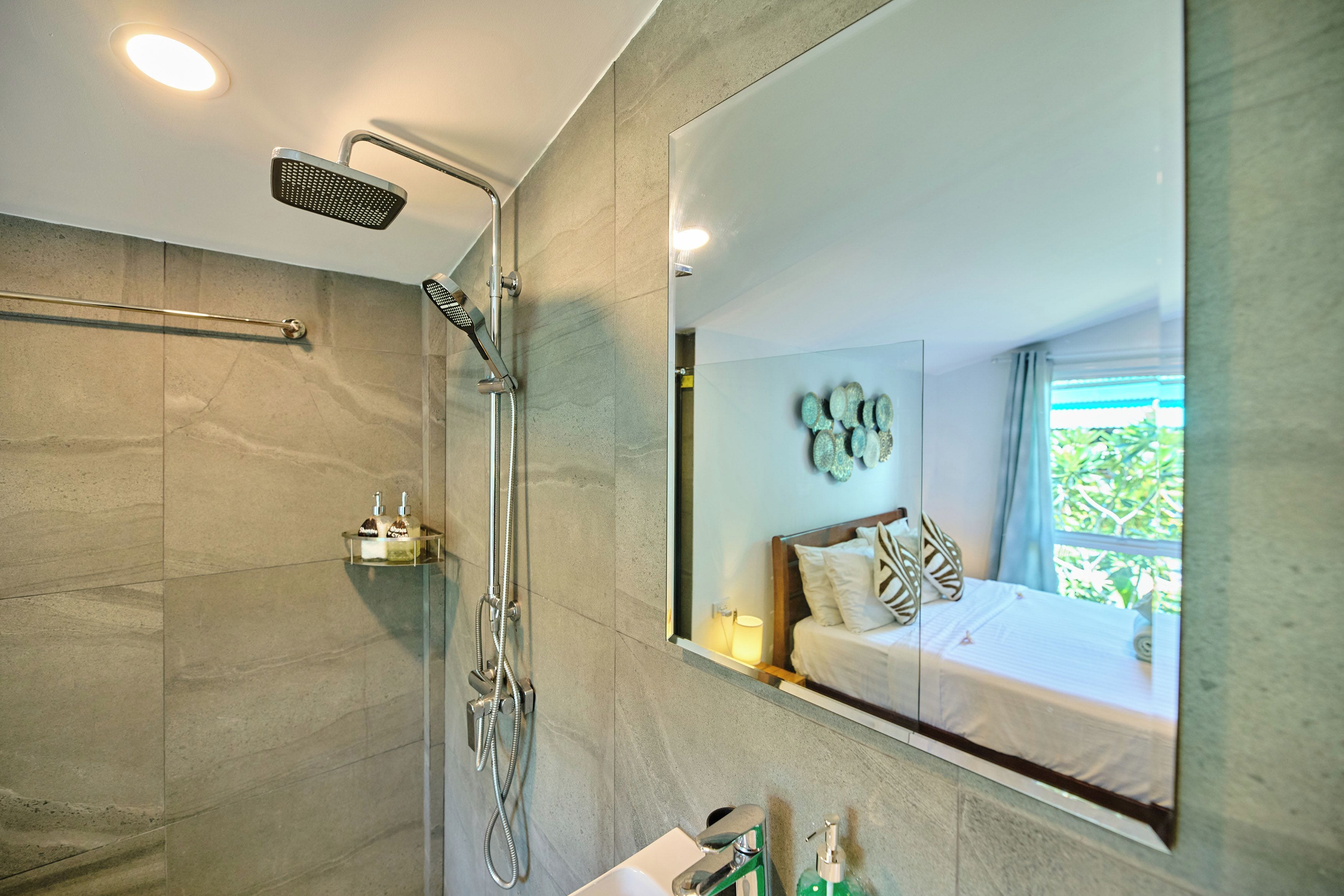 A modern bathroom with a shower, a glass sink, and a large mirror reflecting a bedroom.