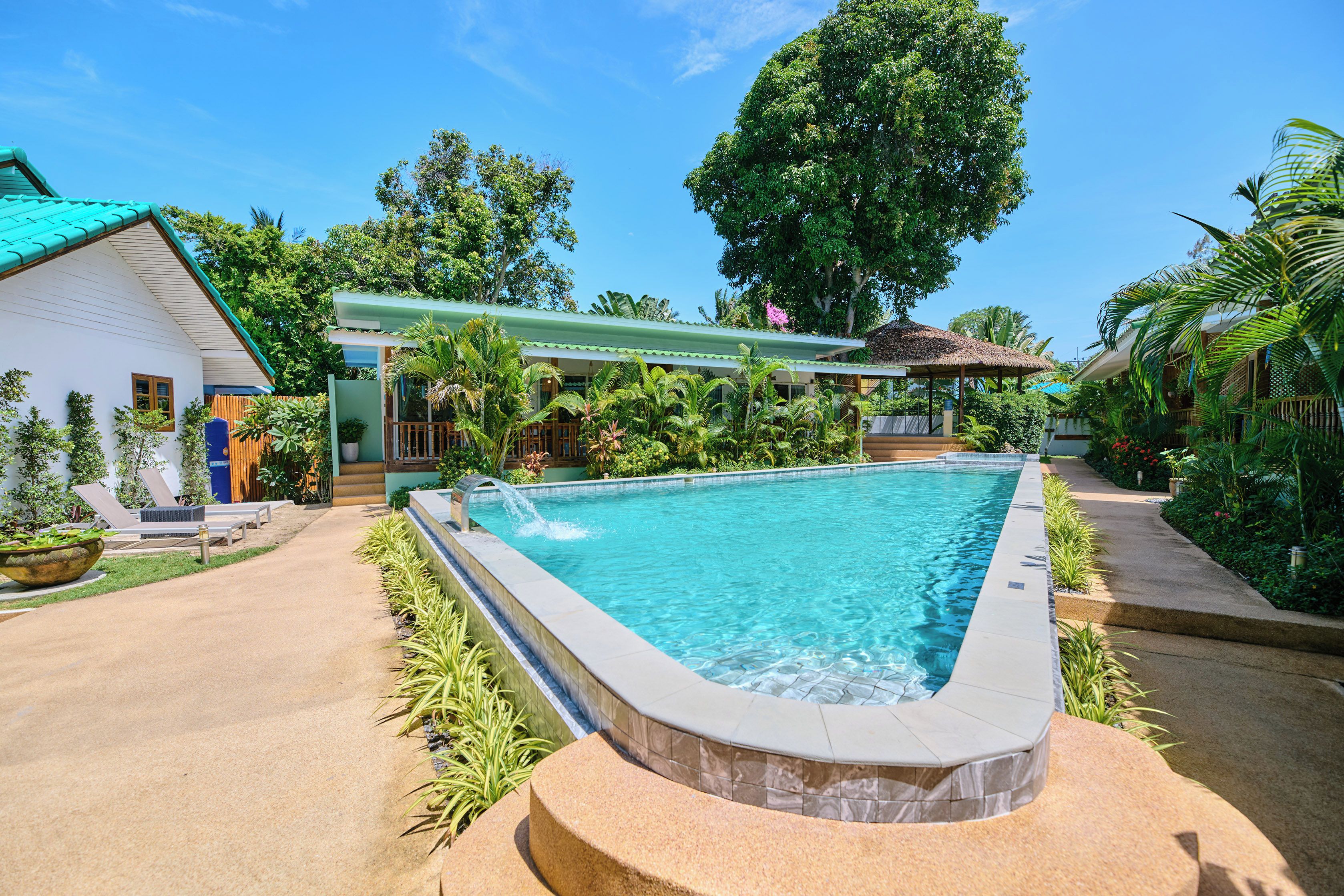 Une piscine rectangulaire bordée de pierres, entourée d’une terrasse en béton. On aperçoit un espace couvert avec un toit végétalisé, ainsi qu’un bâtiment à l’arrière-plan, doté d’une façade blanche et d’un toit bleu.
