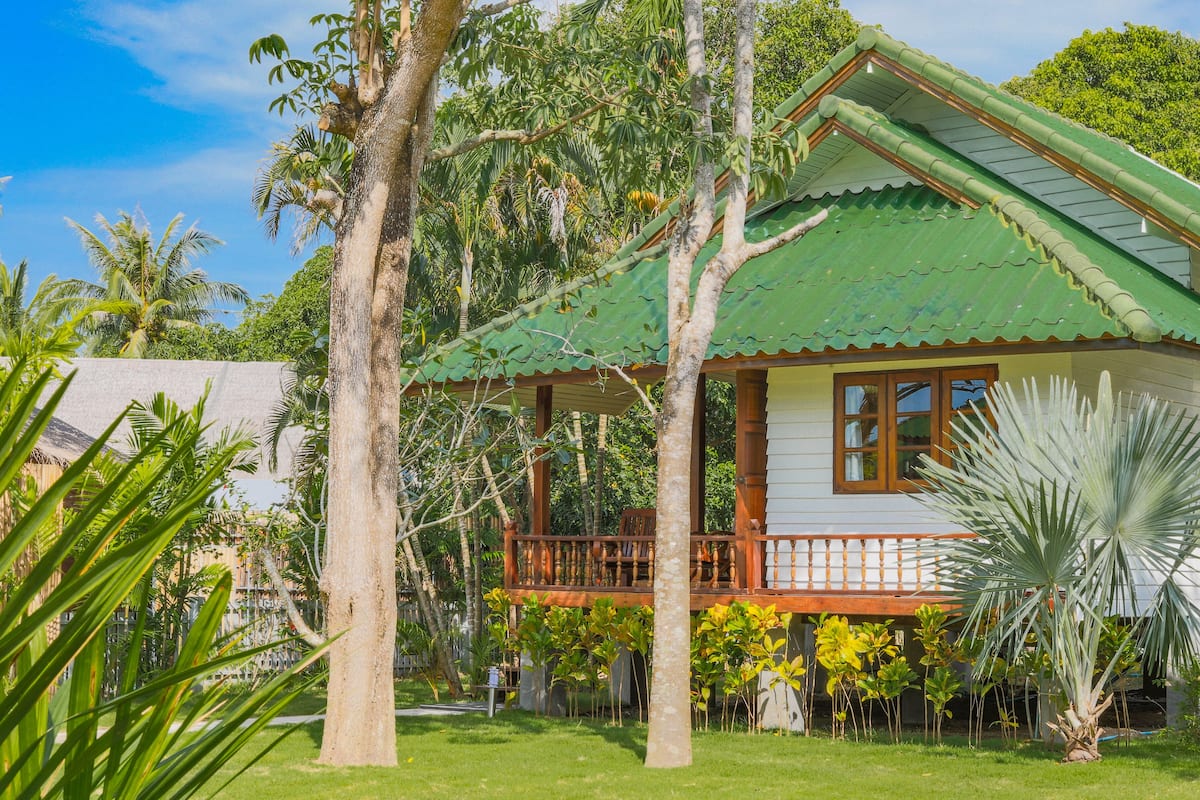 A house with a green roof, wooden balcony, and surrounded by tropical vegetation.