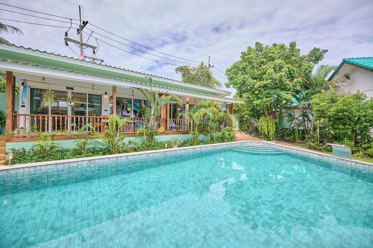 A swimming pool with a tiled edge, surrounded by lush greenery and a building with a green roof.