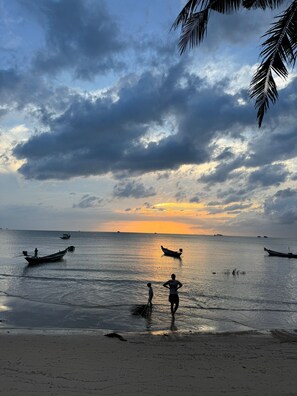 A beach scene with boats on calm water and people standing in the shallow water.