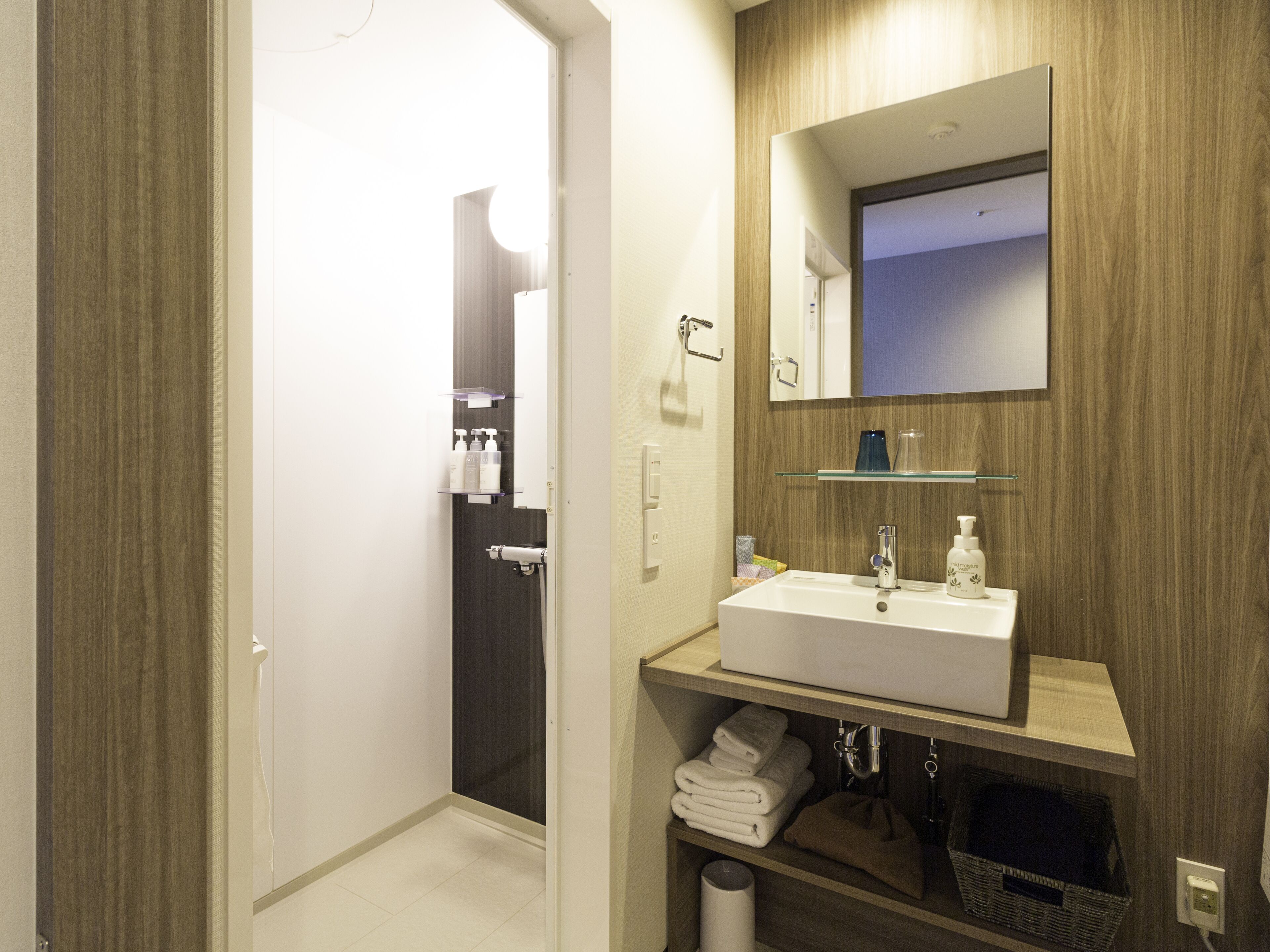 Un baño moderno con lavamanos blanco, espejo y tocador de madera. Hay toallas y artículos de aseo en los estantes.