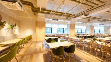 A spacious dining area with wooden tables and chairs, large windows, and exposed ceiling pipes.