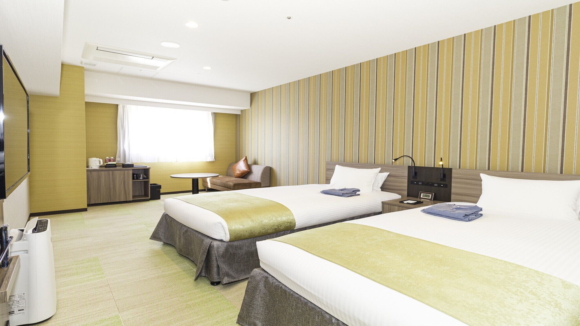 A hotel room with two beds, a small table, a sofa, a TV, and a minibar.