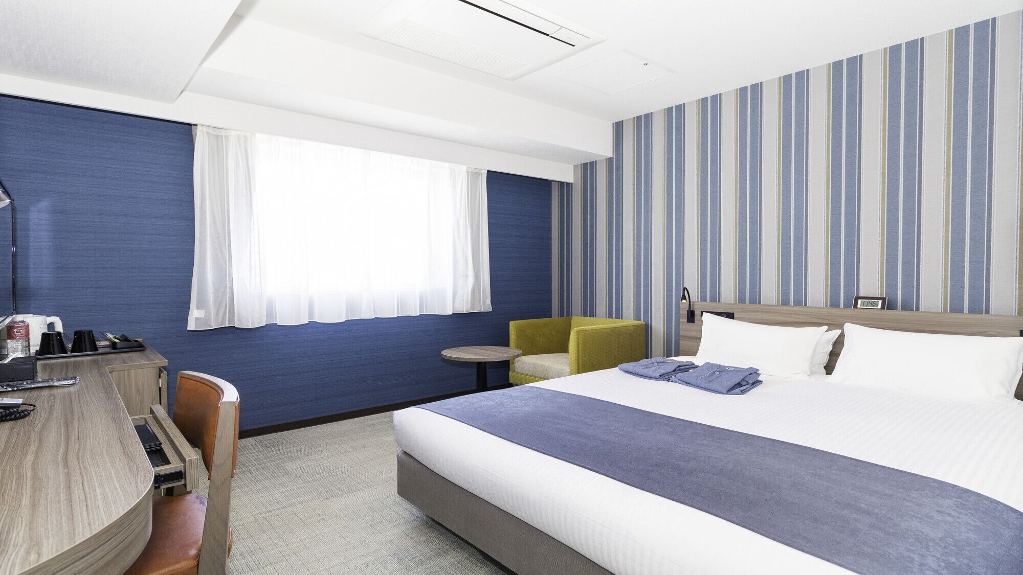 A hotel room with a bed, a desk, a chair, a small table, and a window with curtains.