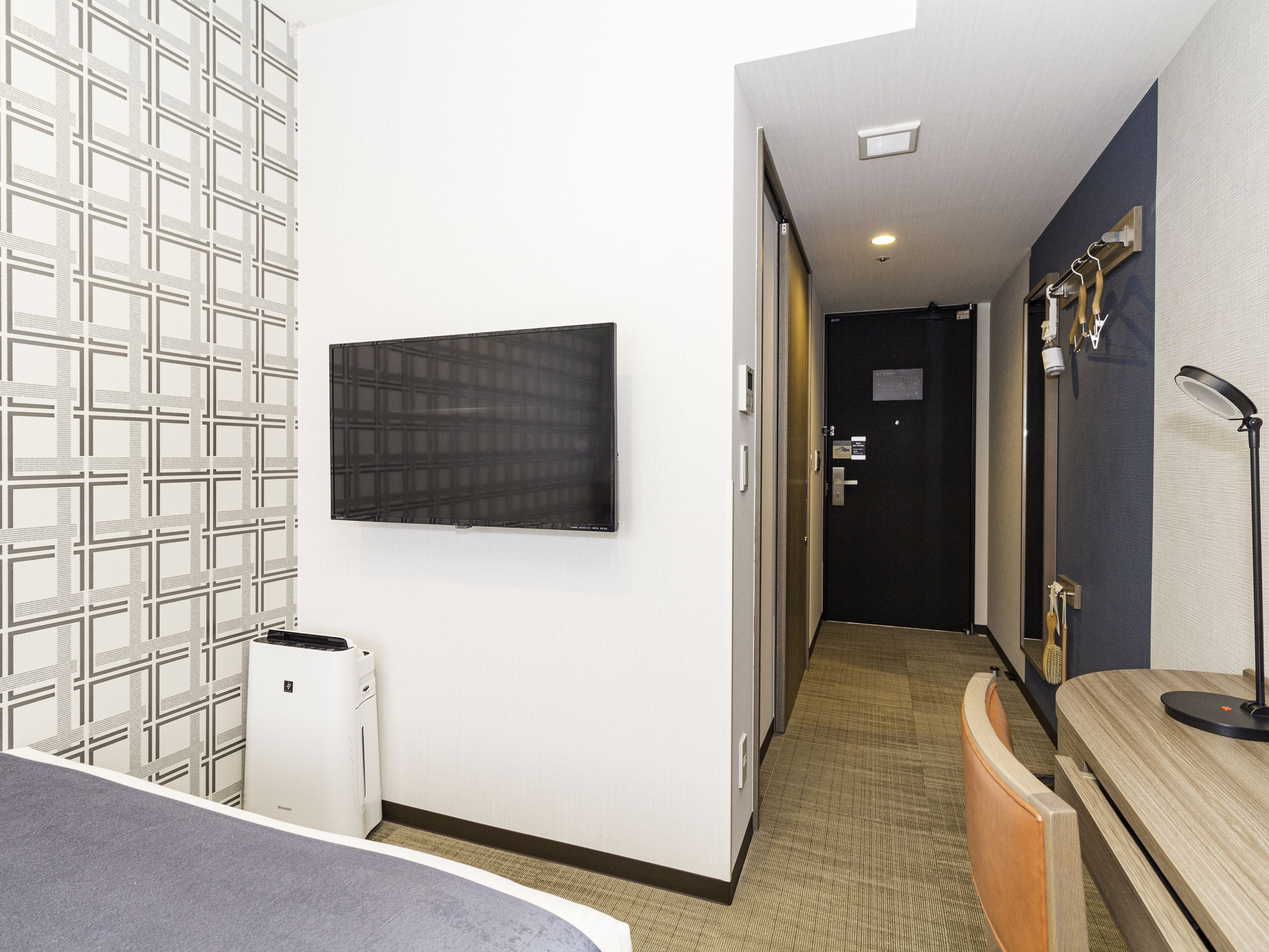 A hotel room with a flat-screen TV mounted on the wall, a white air conditioning unit, a wooden bench, and a mirror with a black frame.