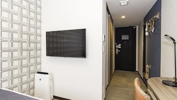 A hotel room with a flat-screen TV mounted on the wall, a white air conditioning unit, a wooden bench, and a mirror with a black frame.