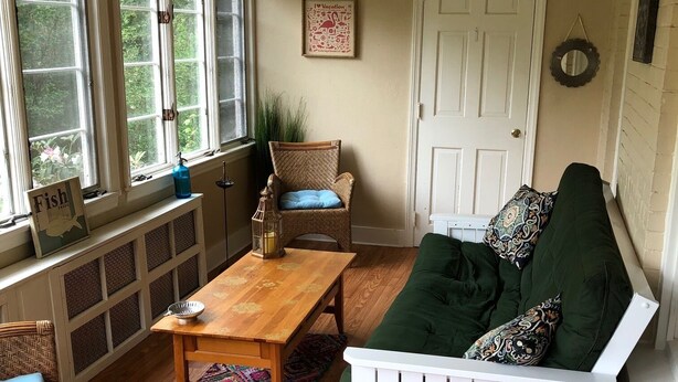 Una habitación acogedora con piso de madera, un banco blanco, un sofá verde y una mesa de madera. Tiene ventanas con cortinas, un ventilador de techo y una puerta.