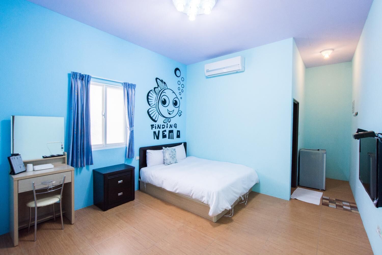 A bedroom with a bed, a desk, a chair, a window with curtains, and a wall decal.