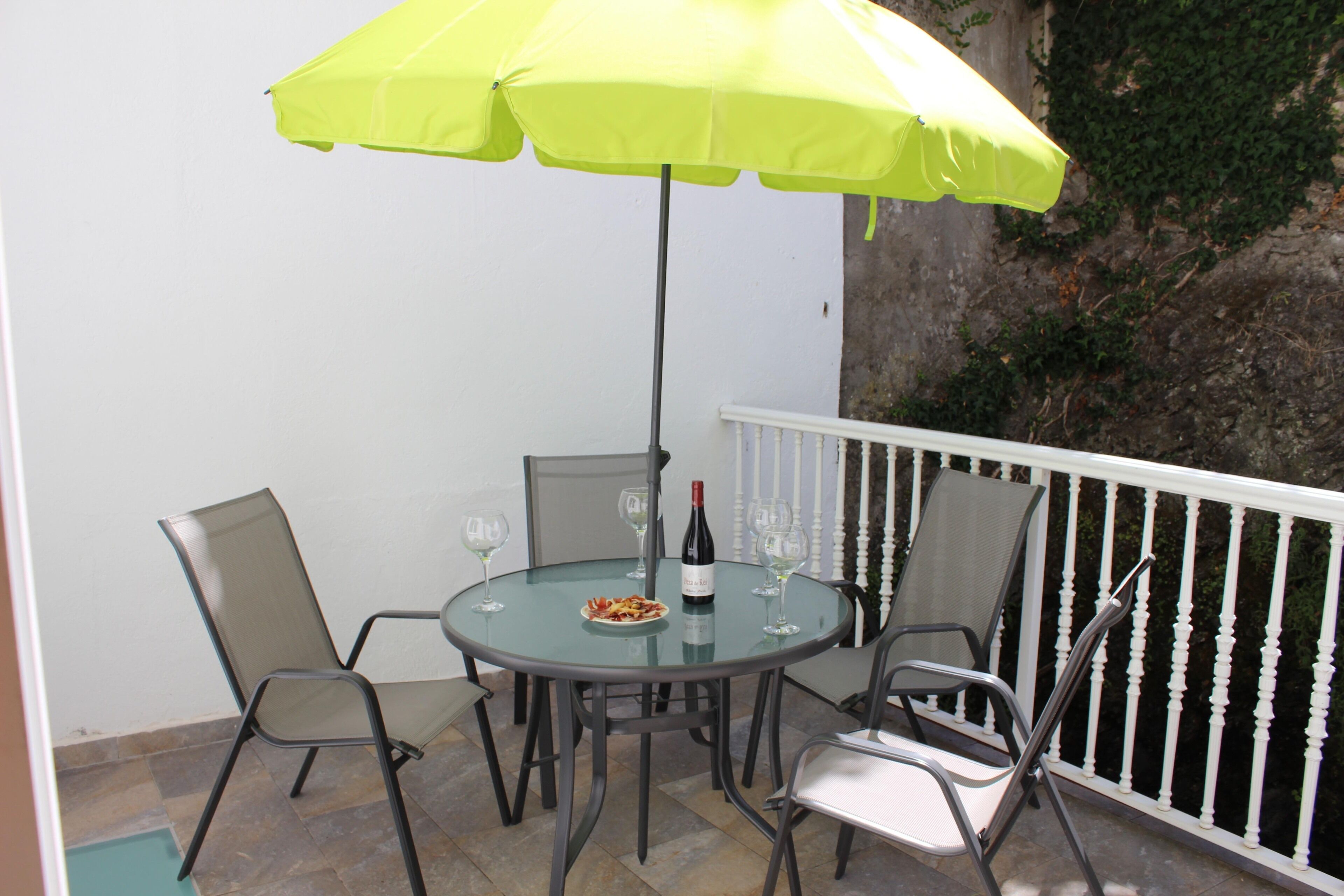 Een ronde glazen tafel met vier stoelen, een fles wijn en twee glaasjes op een balkon met een witte reling.