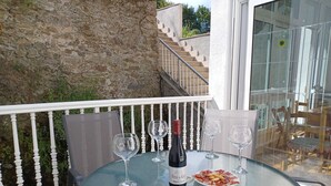 A round glass table with a bottle of wine, two wine glasses, and a plate of food on a balcony with a stone wall and white railing. - Holiday apartment, downtown, seasons (Monforte de Lemos)