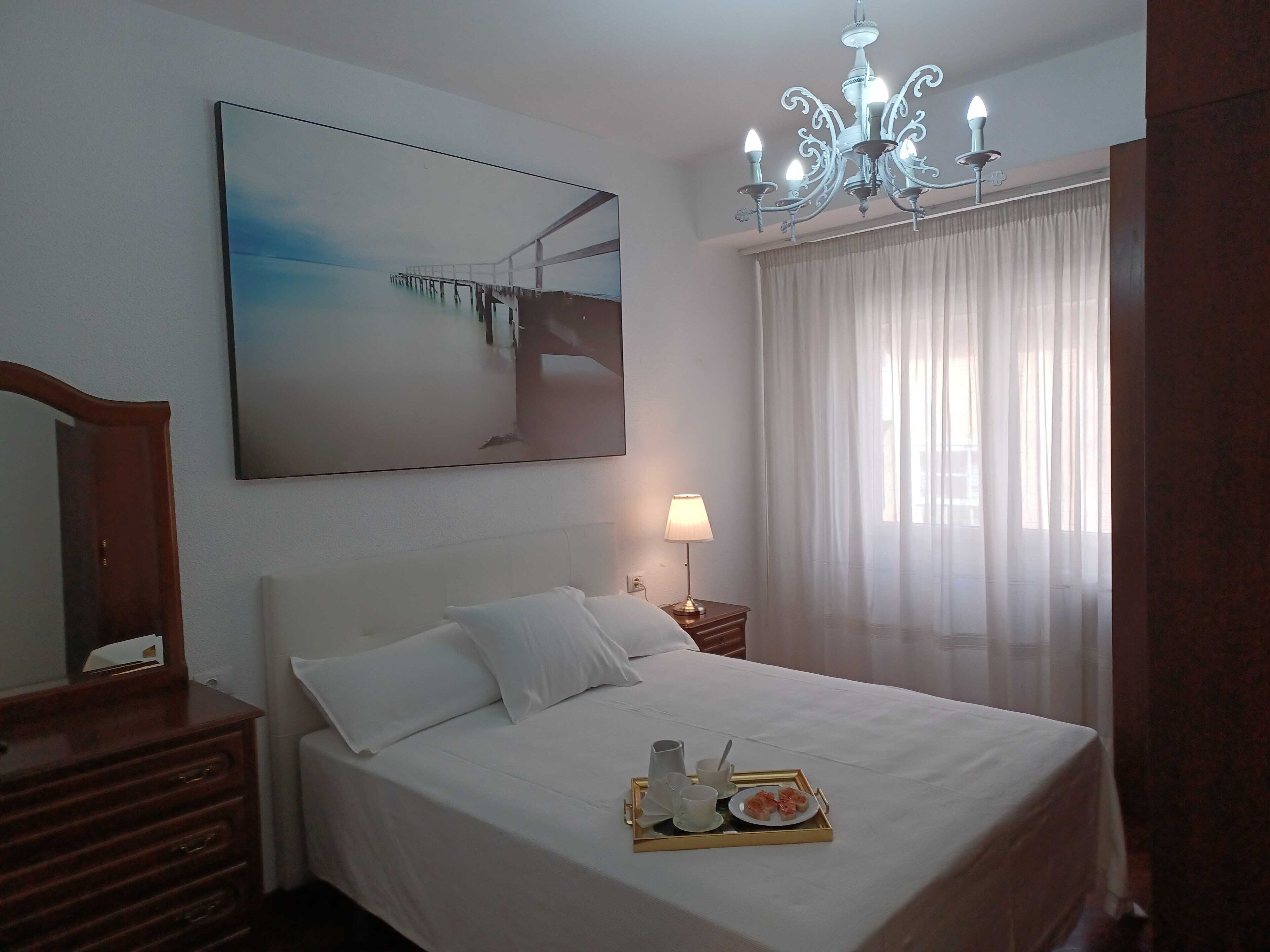 A hotel room with a bed, a bedside table with a lamp, a tray with breakfast items, a large painting on the wall, and a chandelier.