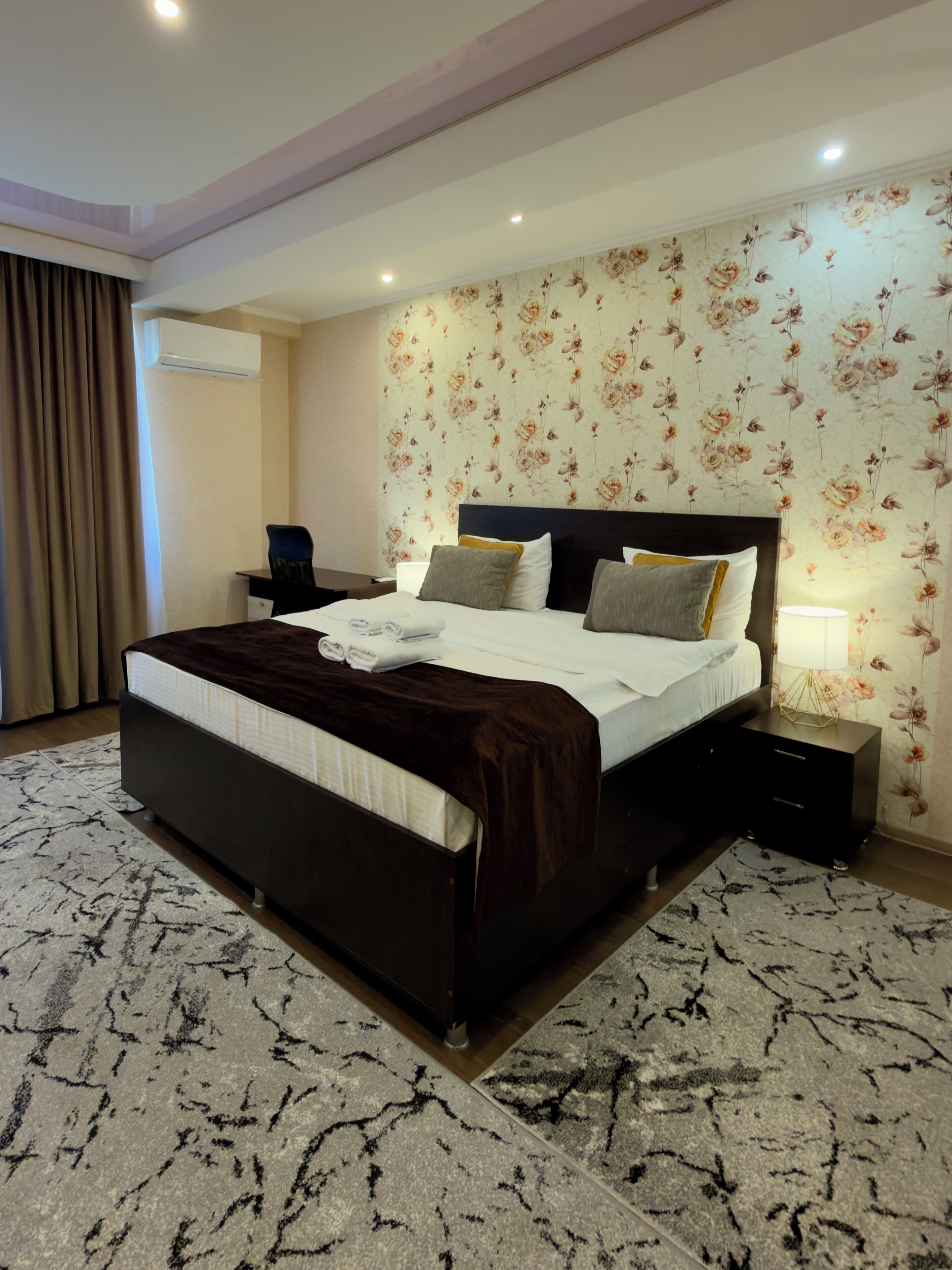 A bedroom with a large bed, a nightstand, a lamp, and a floral wallpaper.