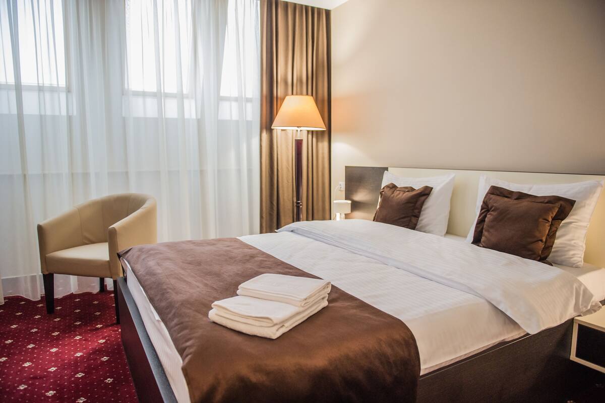 Een hotelkamer met een bed, een stoel, een lamp en een raam met gordijnen.