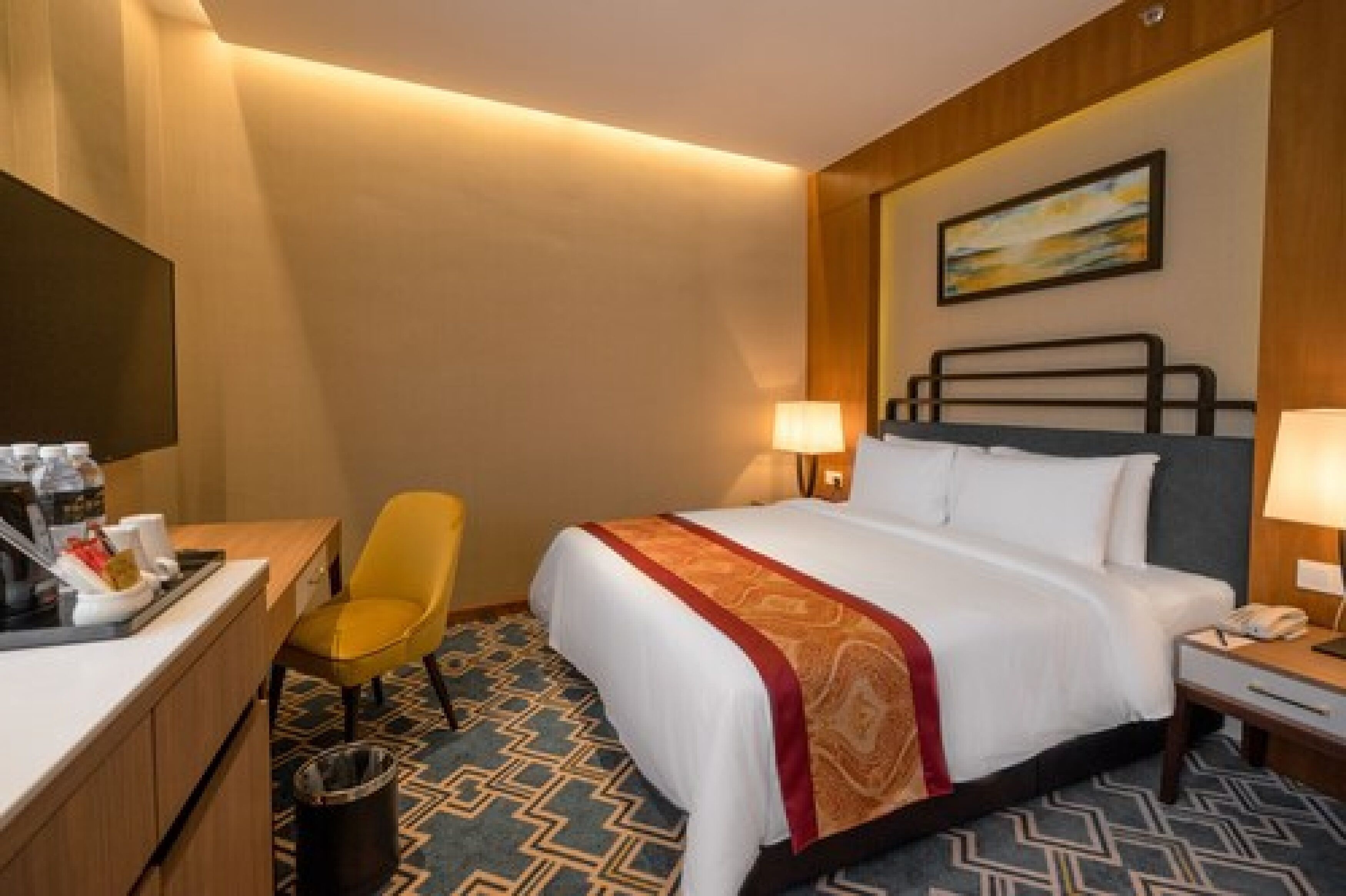 Quarto de hotel com cama, escrivaninha com cafeteira, cadeira, lixeira e um quadro na parede.