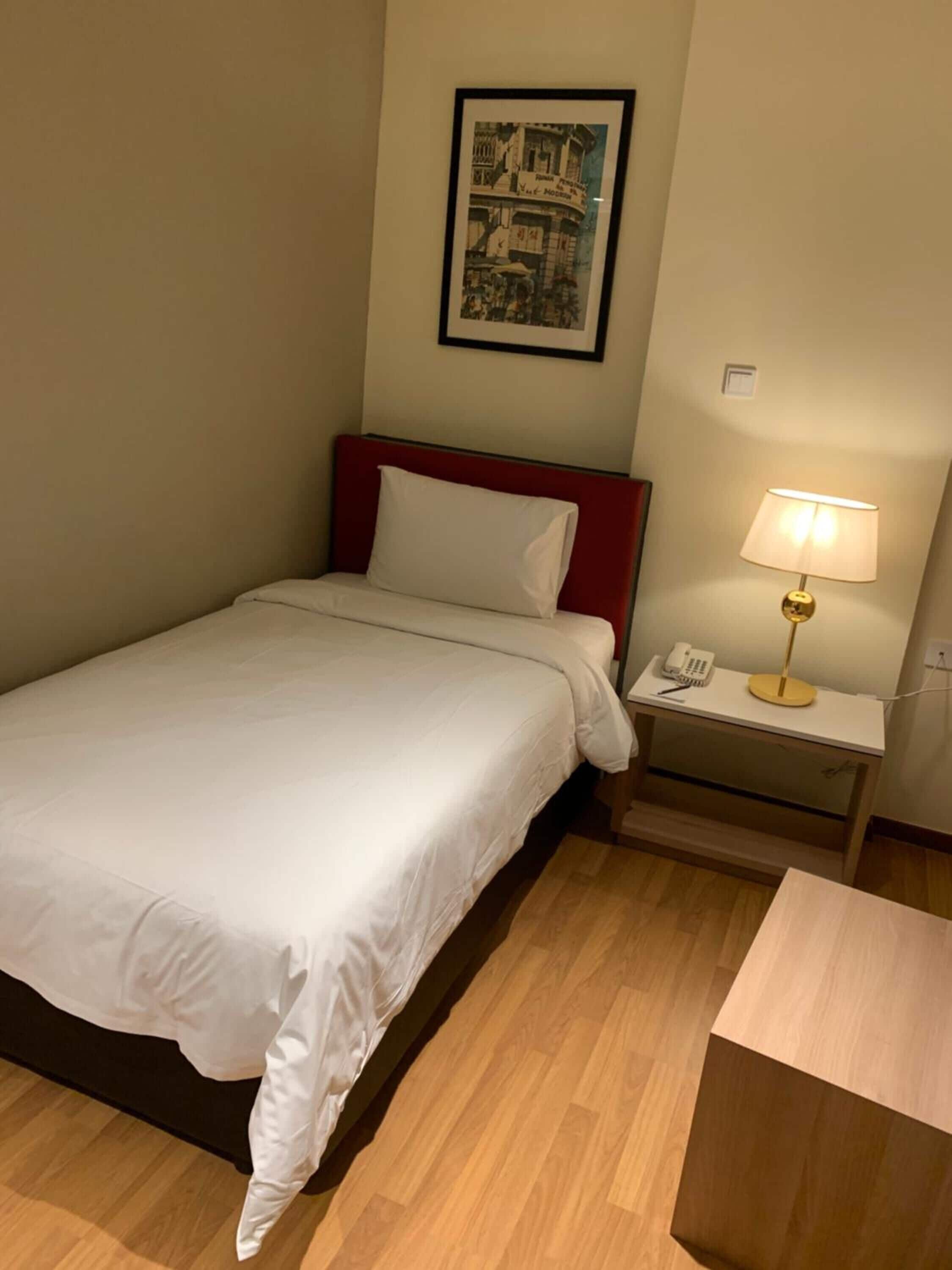A hotel room with a bed, a nightstand with a lamp, a framed picture on the wall, and a wooden nightstand.
