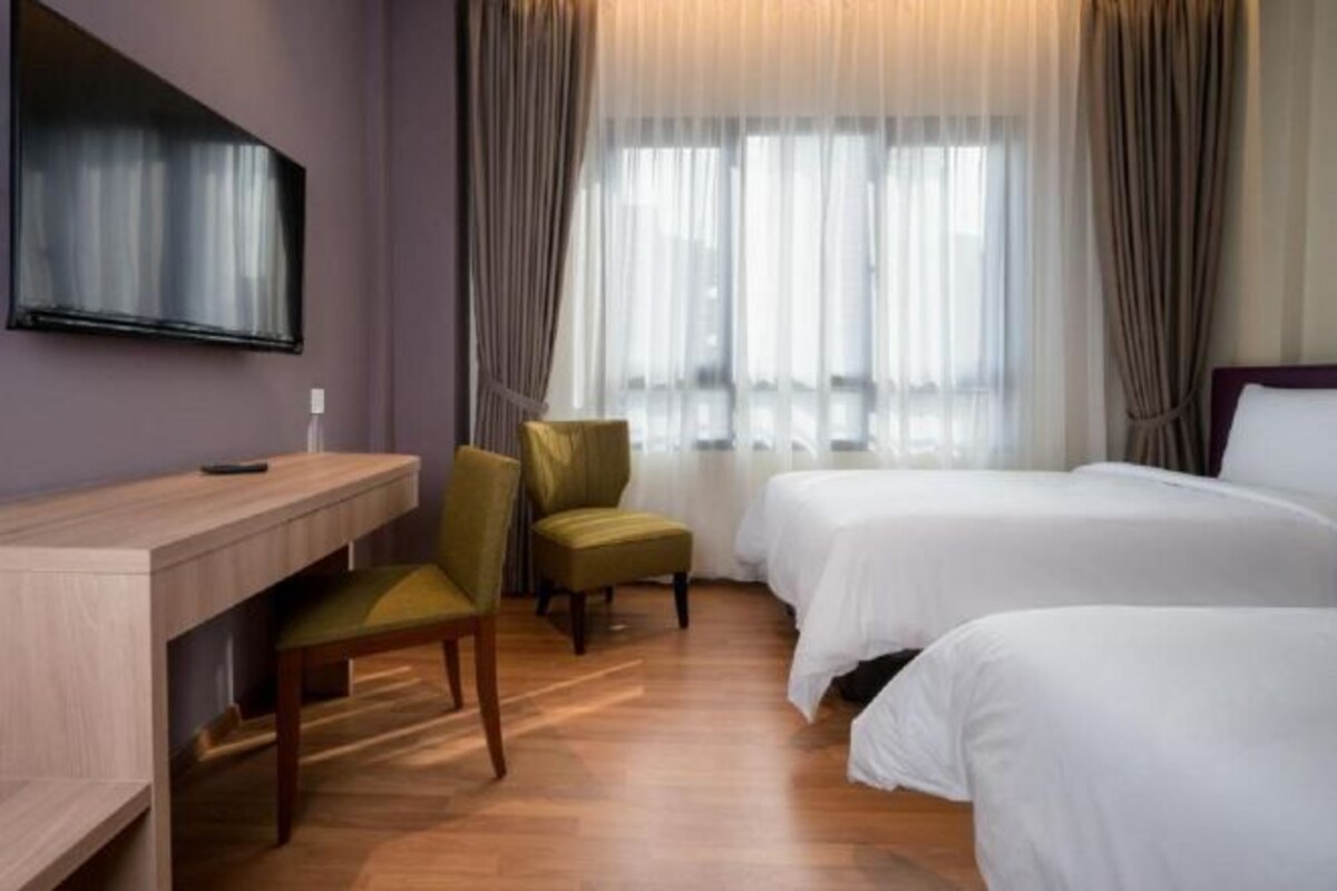 A hotel room with a wooden desk, two single beds, a green armchair, and a large window with sheer curtains.