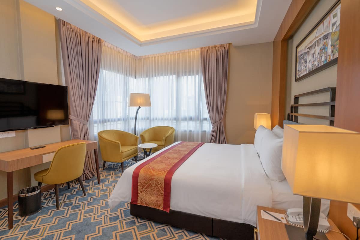 A hotel room with a large bed, a desk with a television, two chairs, a small table, and a lamp.