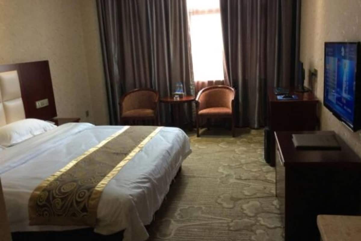 Quarto de hotel com cama, duas cadeiras, uma mesa pequena, televisão e escrivaninha.