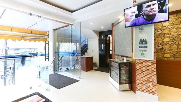 A hotel reception area with a glass door entrance, a staircase, a refrigerator, and a television displaying a news program. - Itsy Hotels Elmas Golden Key (Bengaluru)