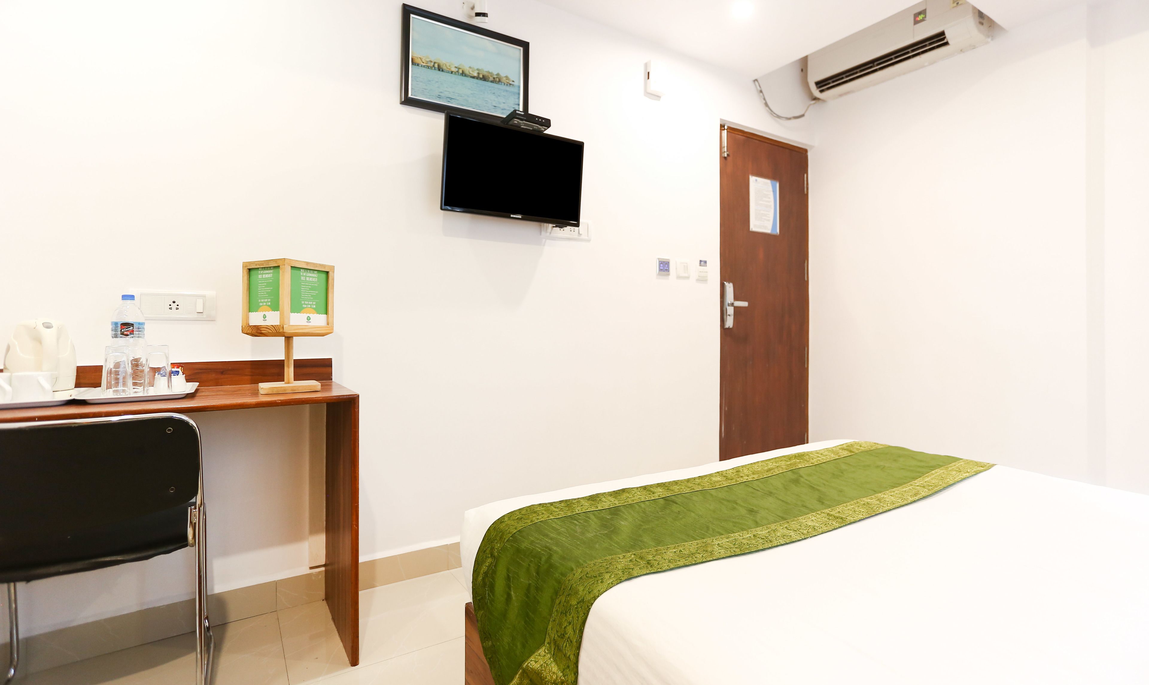 A hotel room with a bed, a desk with a chair, a flat-screen TV mounted on the wall, an air conditioning unit, and a wooden door.