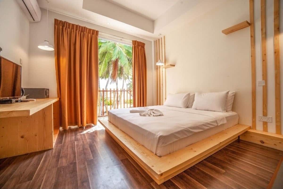 A bedroom with a wooden bed frame, a desk with a computer, and a view of palm trees through large windows.
