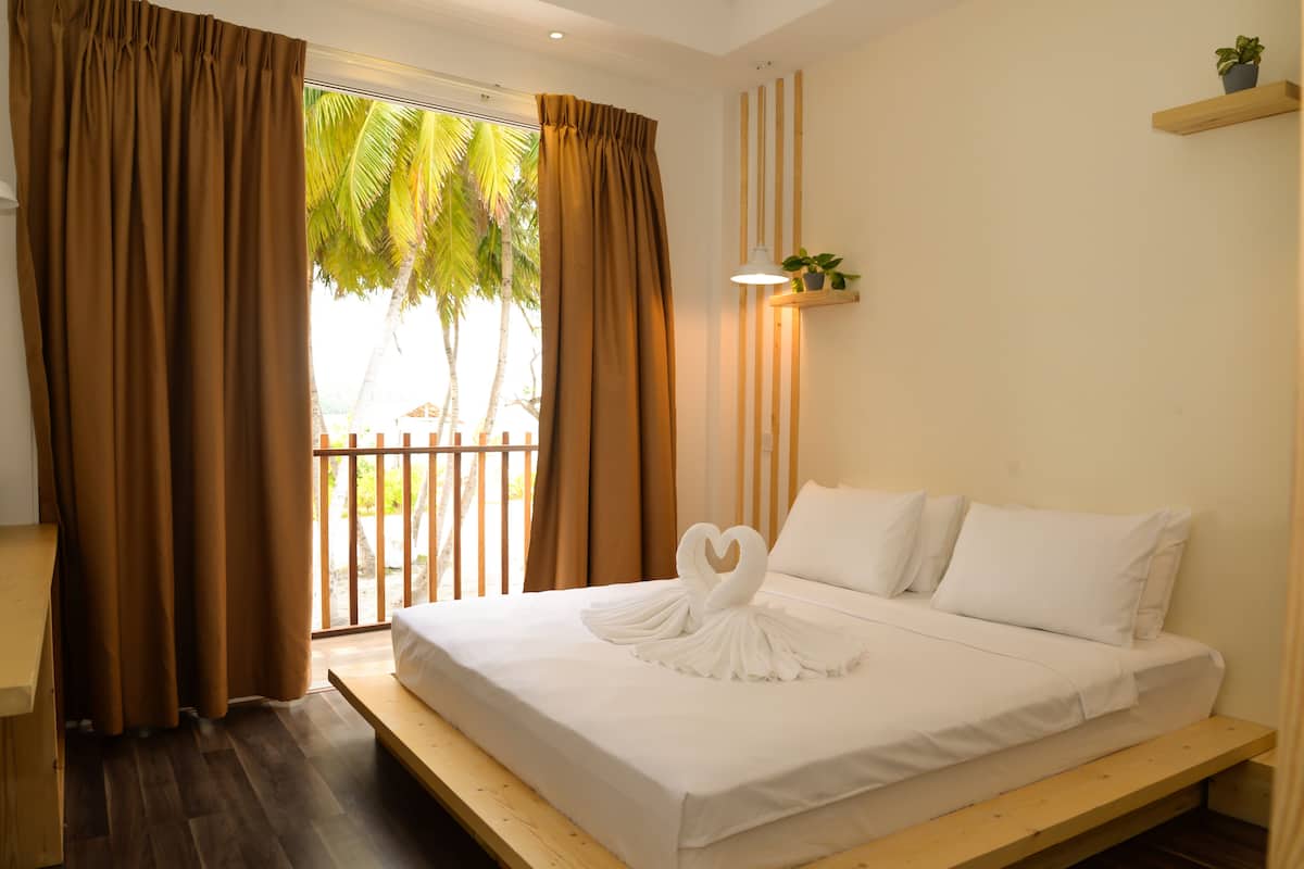 A bedroom with a bed, curtains, a wooden bench, a potted plant, and a view of palm trees.