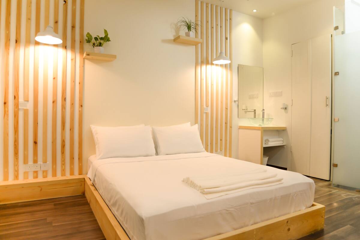 A neatly arranged bedroom with a wooden bed frame, a bedside table with a mirror, and a small shelf with a plant.
