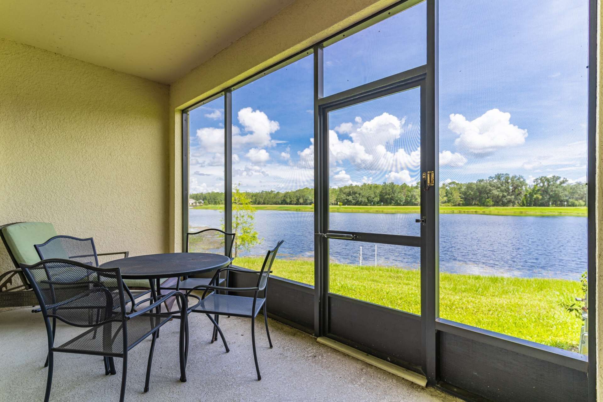 A covered patio with a table and chairs, overlooking a lake with grassy banks and trees.