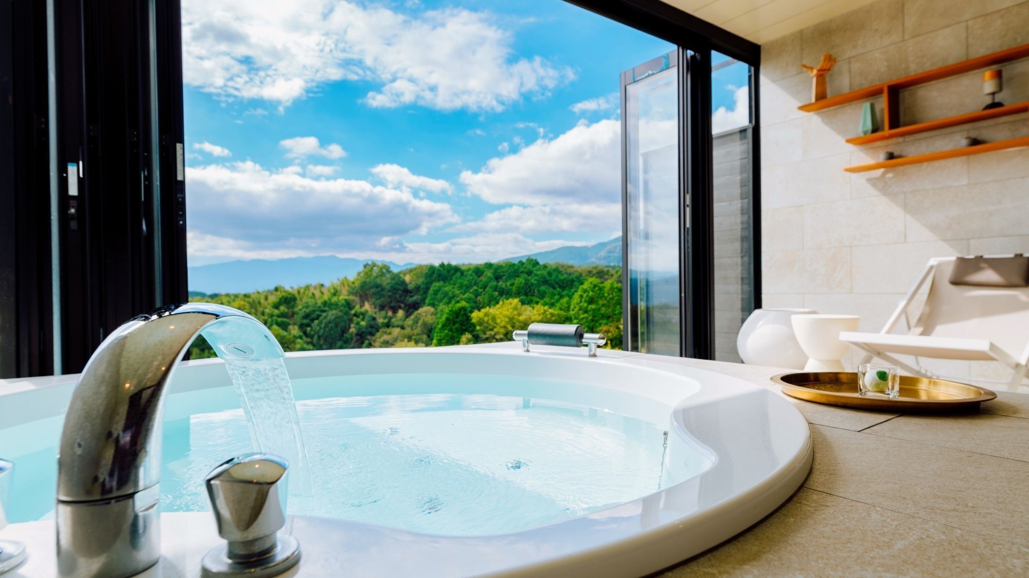 A hot tub with a waterfall feature, a glass door leading to a balcony with a view of a forest and mountains, and a white chair on the balcony.
