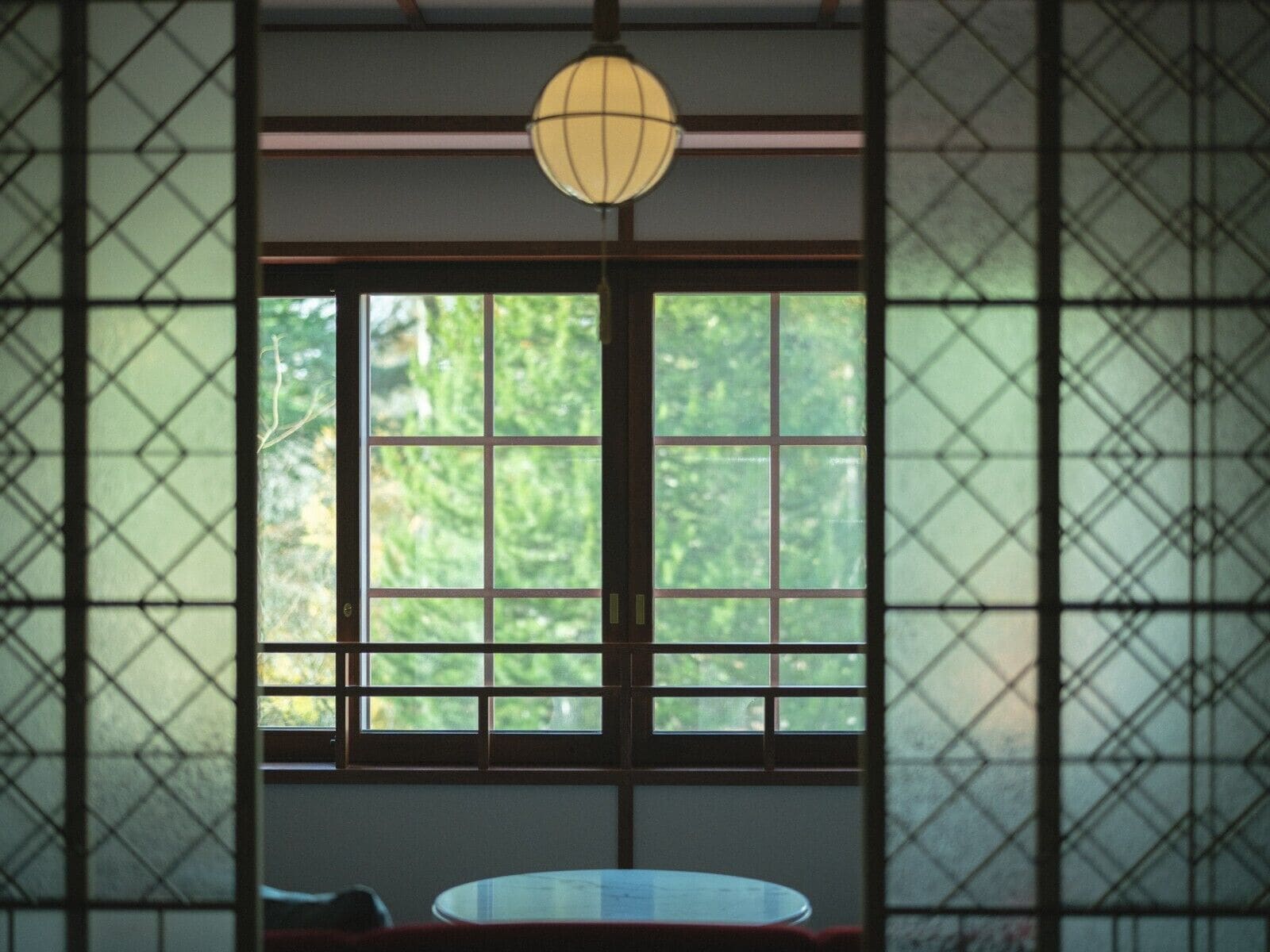 A room with a large window, a round table, and a hanging light fixture.