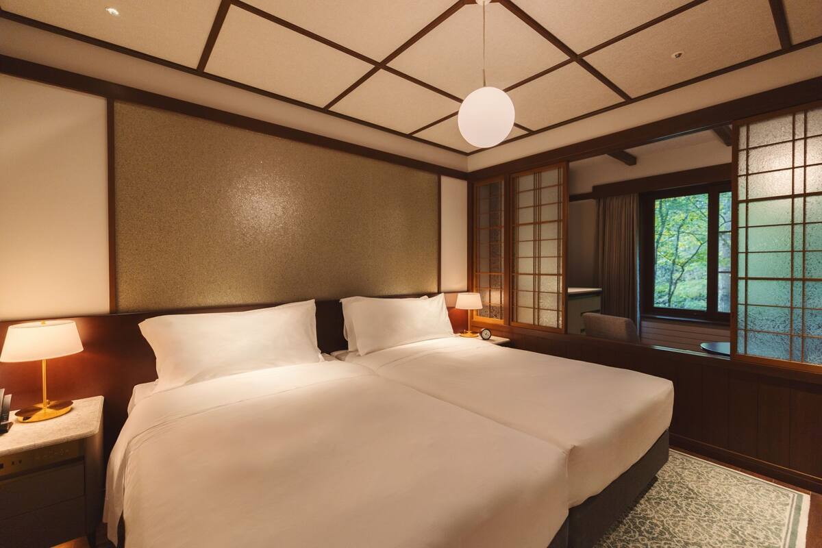 A hotel room with a large bed, two bedside lamps, a wooden headboard, a view of greenery through a window, and a traditional Japanese-style wall panel.