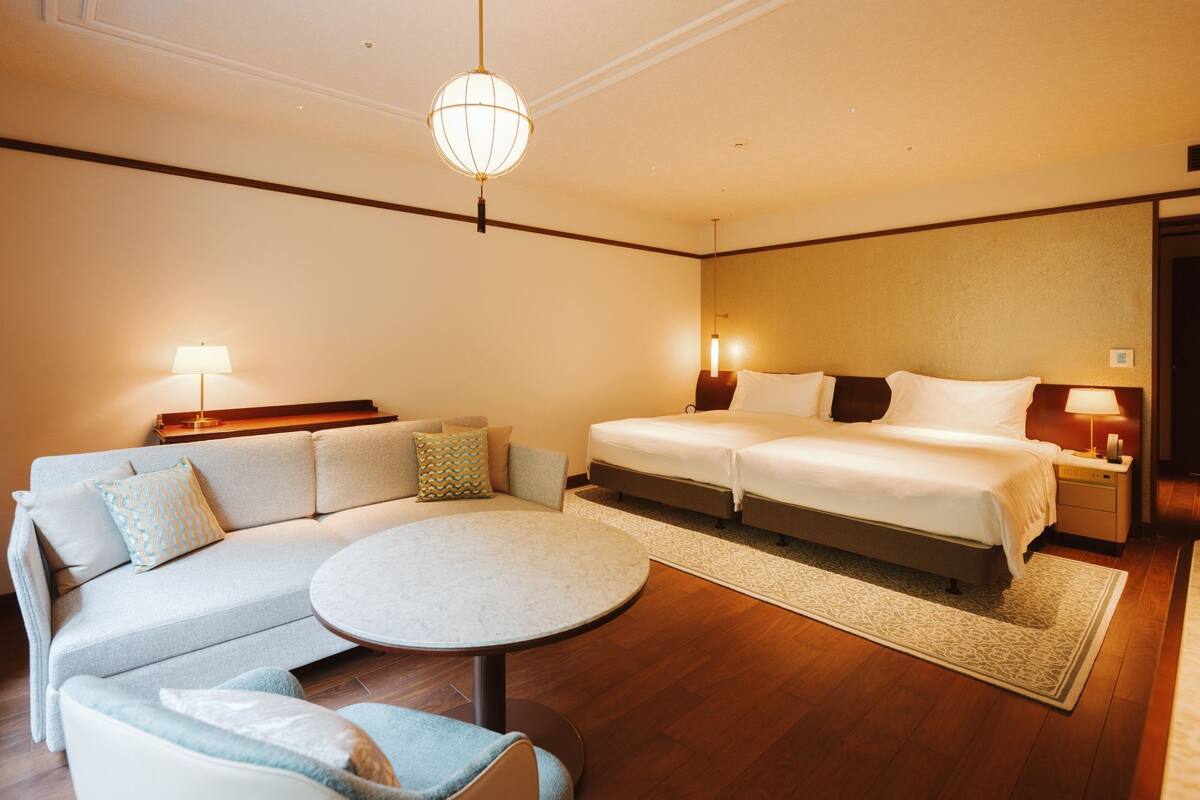 A hotel room with a sofa, a round table, a bed with two pillows, and a nightstand with a lamp.