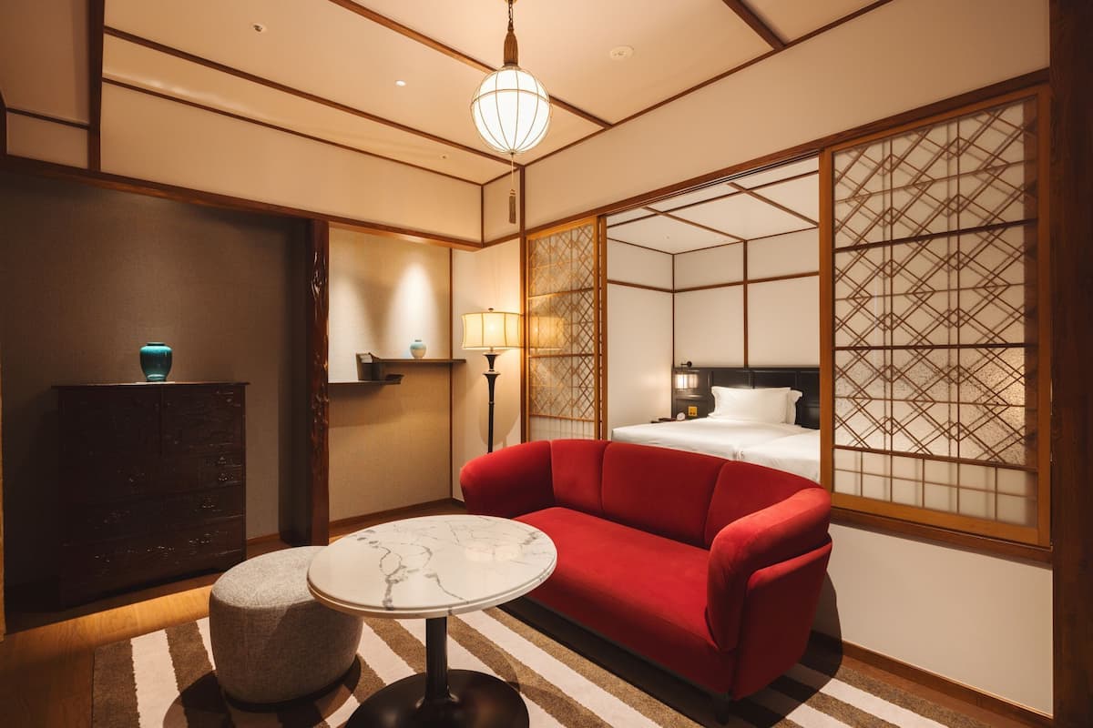 A hotel room with a sofa, a small round table, a lamp, a decorative vase, and a bed with a headboard.
