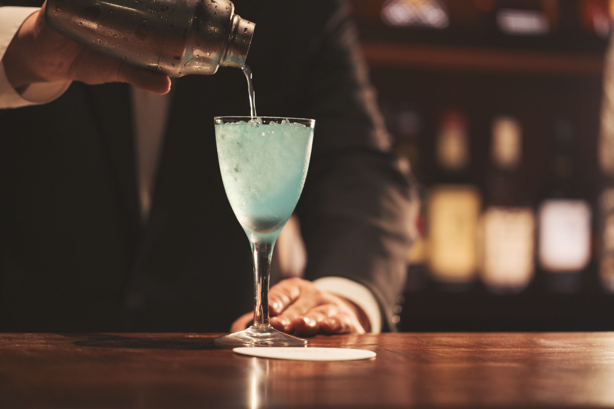A person pouring a blue liquid into a martini glass on a bar.