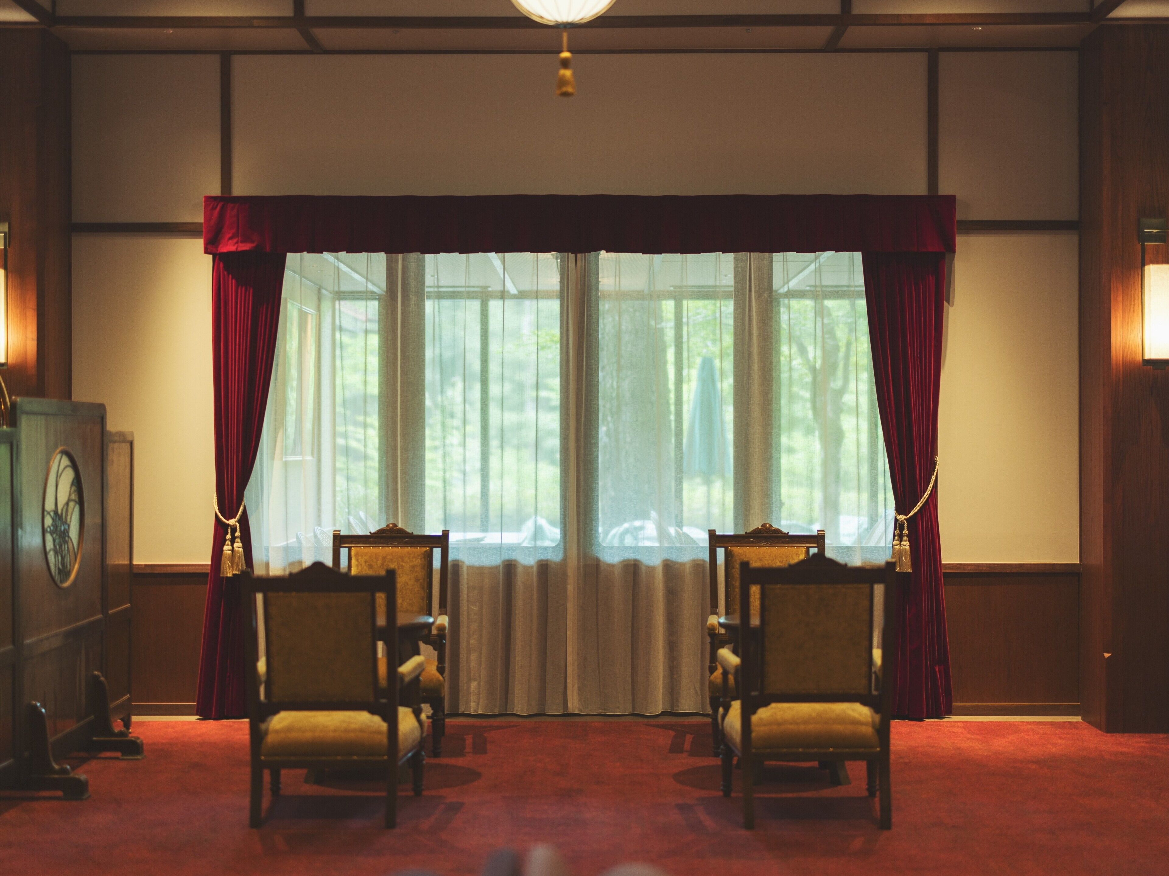 A room with two wooden chairs, a red carpet, a large window with curtains, and a wooden wall.
