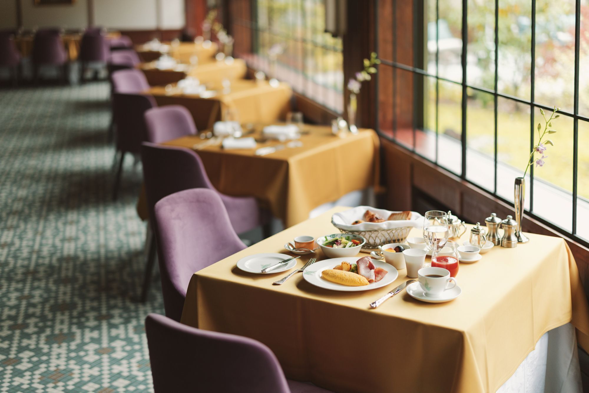 A restaurant with tables set for dining, featuring a patterned carpet and large windows.