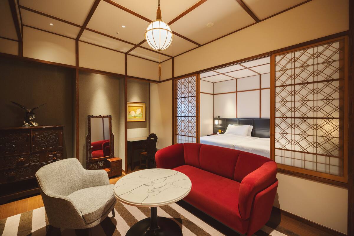 A hotel room with a red sofa, a small round table, a wooden dresser, and a traditional Japanese sliding door.