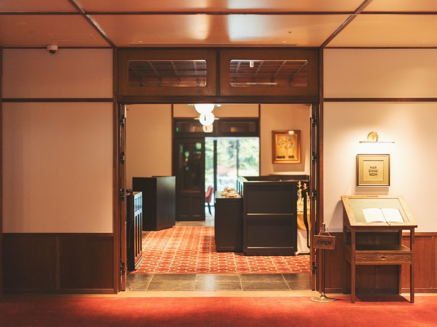 Un vestíbulo de hotel con alfombra roja, un mostrador de recepción y un cartel enmarcado que indica el comedor principal.