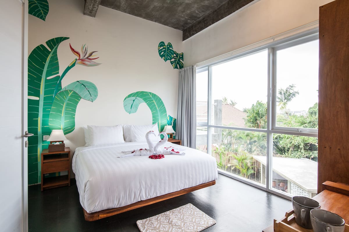 A modern bedroom with a large bed, a view of the outdoors, and tropical wall art.