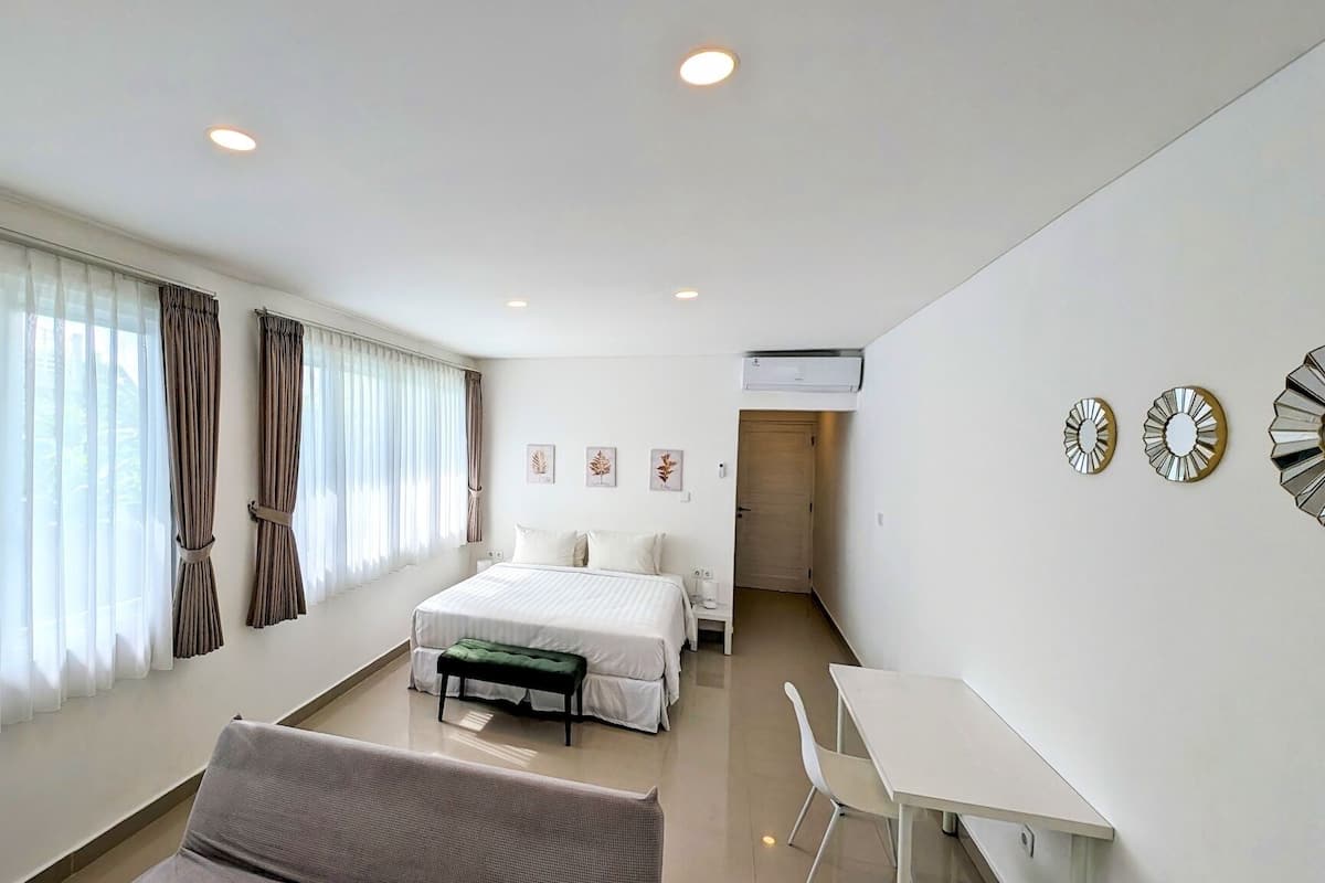 A modern hotel room with a bed, a desk, and a chair. There are curtains on the windows, and an air conditioning unit is visible on the wall.