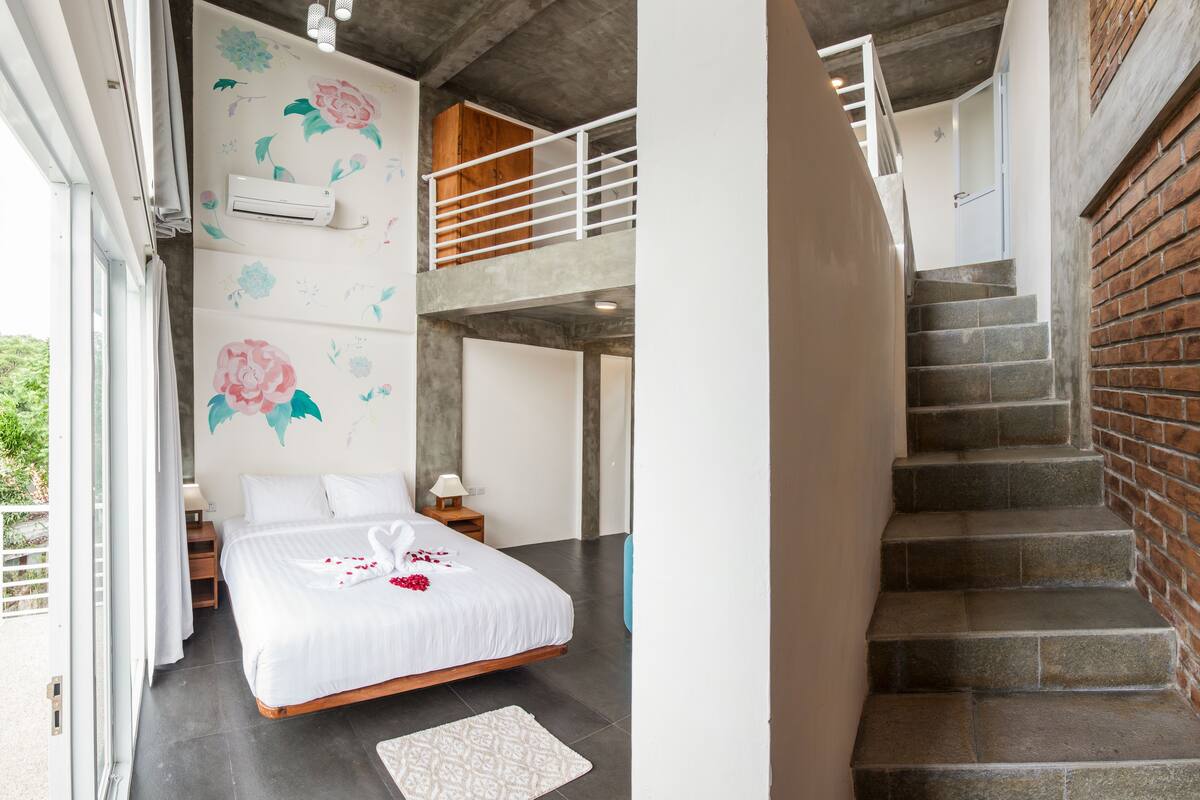 A bedroom with a large bed, a wooden ceiling, a staircase, and a brick wall.