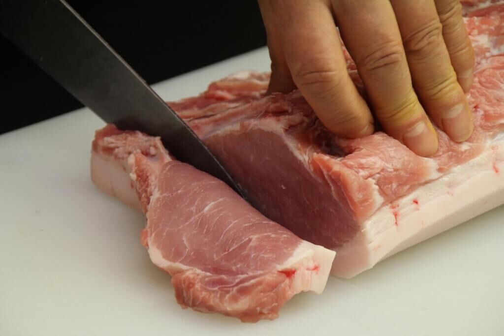 Uma mão segurando uma faca cortando um pedaço de carne crua em uma tábua de corte branca.