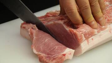 Uma mão segurando uma faca cortando um pedaço de carne crua em uma tábua de corte branca.