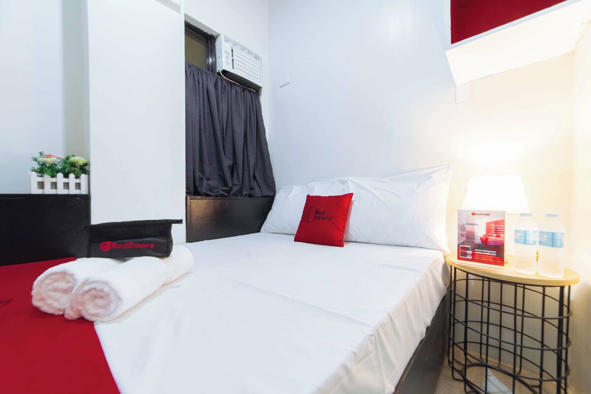 Ein Hotelzimmer mit einem ordentlich bezogenen Bett, einem roten Kissen, einem kleinen Tisch mit Lampe und Wasserflaschen sowie einer weißen Wand mit Fenster und Klimaanlage.