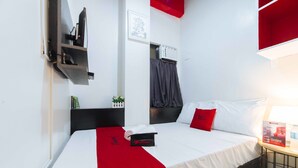 Une chambre d’hôtel avec un lit, un plaid rouge, une table de chevet noire, un oreiller rouge, un mur blanc, un tableau encadré et une fenêtre avec des rideaux.