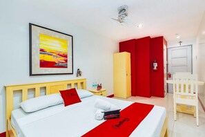 A hotel room with a bed, a red and white bedspread, a framed abstract painting, a wooden nightstand, a red door, and a white chair. - RedDoorz near Cebu Metropolitan Cathedral (Cebu City)
